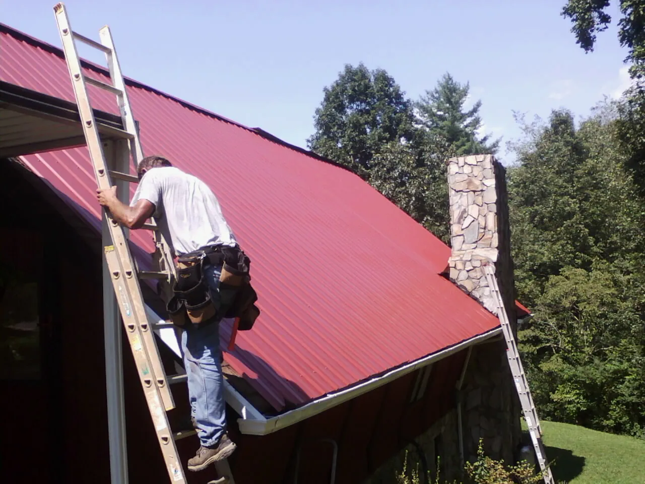 Roofer on a ladder beside a red metal roof and stone chimney — Soffit & Fascia Repair by Roofing Brothers in Wyatt